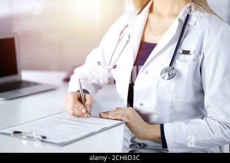 Unbekannte Ärztin verschreibt ihrem Patienten Medikamente, indem sie eine Zwischenablage benutzt, während sie am Schreibtisch in ihrem sonnigen Schrank sitzt. Weiblich Stockfoto