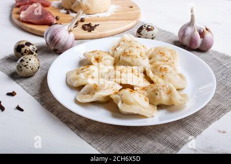 Knödel auf einem Teller mit Zutaten (Fleisch, Teig, Gewürze) auf einem Leinentischtuch auf weißem Holzhintergrund. Nahaufnahme. Stockfoto