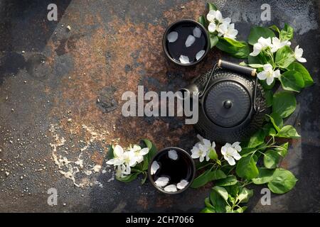 Schwarze Keramik Teekanne und Tasse mit heißem Tee von Frühlingsblüten Apfelzweige über dunkle Textur rustikalen Eisen Hintergrund dekoriert. Flach legen Frühling Tee c Stockfoto