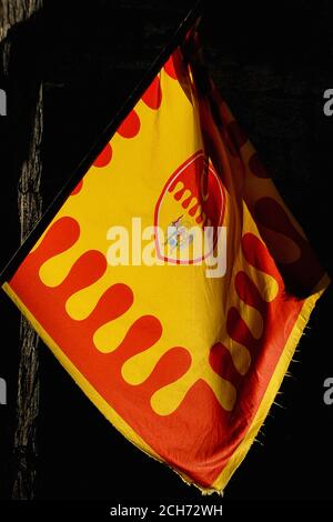 Leuchtende gelbe und rote Flagge der Contrada (historisches Viertel) von Sant’Agnolo leuchtet in der Abendsonne in Volterra, Toskana, Italien. Das Mittelalter kontrahierte die Bewohner jedes Gebietes, um sich gegenseitig zu unterstützen. Sant’Agnolo oder Sant’Angelo gehörte zu den 1779 abgeschafften Kontrachten, doch der heutige toskanische Tourismusboom hat ihre Wiederbelebung ausgelöst. Contrade-Flaggen wurden in einer Prozession getragen und die ursprüngliche Sant’Agnolo-Flagge wurde 1236 in die Schlacht getragen. Diese moderne Version, die vor Volterras jährlichem Mittelalterfest abgebildet wurde, enthält das abnehmende Mond-Emblem der contrada und eine Darstellung des Heiligen Erzengels Michael. Stockfoto