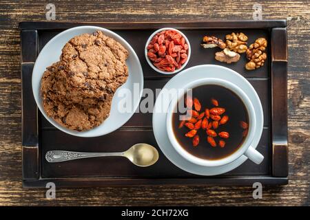 Weiße Tasse frischen Goji-Kaffee am Morgen mit Keksen auf Holztablett auf dem Tisch, Draufsicht, Nahaufnahme. Schwarzer heißer Kaffee mit ganzen roten Goji-Beeren Stockfoto