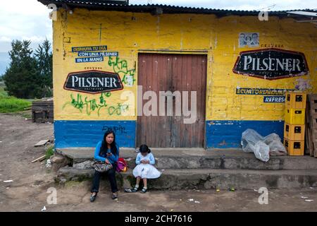Eine Dame und ein Mädchen in einem hübschen Kleid sitzen vor einem bunten Geschäft am Stadtrand von Quinche in Ecuador. Stockfoto