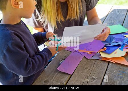 Kleines Kind im Alter von 3 Jahren lernen, Papier mit einer Schere zu schneiden Draußen auf einem Tisch mit buntem Kunstpapier sitzen Carmarthenshire Wales Großbritannien KATHY DEWITT Stockfoto