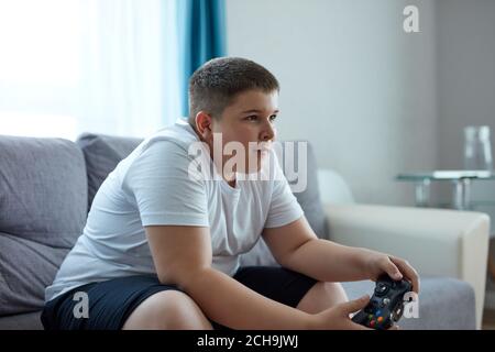 Keen kaukasischen Jungen spielen Videospiele, übergewichtigen Jungen sitzen auf dem Sofa allein, hat Probleme mit der Ernährung, führen ungesunden Lebensstil. Faulheit Konzept Stockfoto