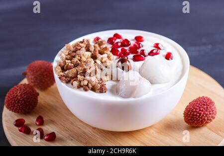 griechischer Joghurt mit Litschi, Granatapfelkernen und Granola in einem weißen Teller auf schwarzem Holzhintergrund, Nahaufnahme. Stockfoto