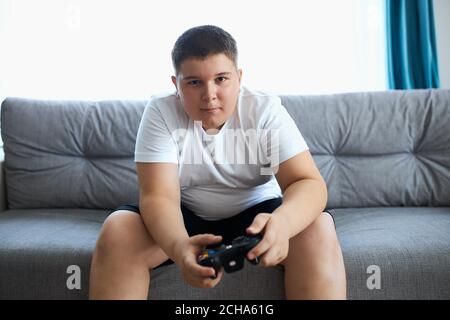 Keen kaukasischen Jungen spielen Videospiele, übergewichtigen Jungen sitzen auf dem Sofa allein, hat Probleme mit der Ernährung, führen ungesunden Lebensstil. Faulheit Konzept Stockfoto