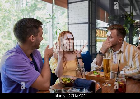 Kleine Aufkleber auf der Stirn. Freunde, die Spaß im Restaurant haben, kaukasische junge Leute spielen ein Hedbanz-Spiel, lachen Stockfoto