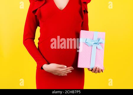 Zugeschnittenes Bild von rosa gefleckte Geschenkbox in der Hand der schwangeren Frau gegen ihren Bauch auf gelbem Hintergrund. Zukünftige Mutter in rotem Kleid. Warten auf ein Baby Stockfoto