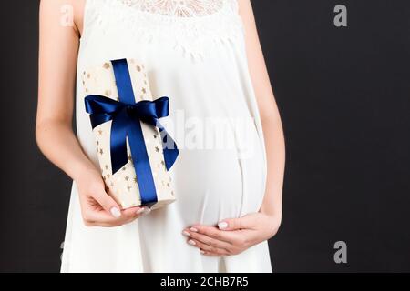 Zugeschnittenes Bild von schwangeren Frau in weißem Kleid hält eine Geschenkbox und berühren ihren Bauch auf schwarzem Hintergrund. Erwartet einen kleinen Jungen. Speicherplatz kopieren. Stockfoto
