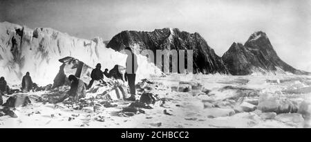 Die Party lebte auf Elephant Island, bis Sir Ernest Shackleton sie rettete, nachdem sie aus dem Eisloch vertrieben worden waren. Das Packeis wird gesehen, wie es die Bucht erstickt, während im Hintergrund die Gletscher zu sehen sind, von denen Lawinen drohten, die Bucht zu füllen. Im Vordergrund sind Mitglieder der Expedition zu sehen, die die Gentoo-Pinguine, die ihr Hauptfutter für 4 Monate gebildet haben, enthäuten. Stockfoto