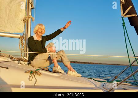 Glückliche ältere Frau sitzt auf der Seite des Segelbootes oder Yacht Deck schwimmend in der ruhigen blauen Meer, wegschauen und winken zu jemandem Stockfoto