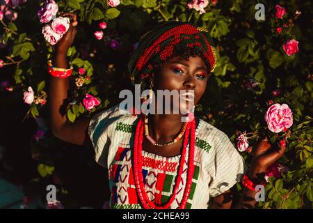 Zugeschnittenes Bild der afrikanischen weiblichen Modelin Nationalkostüm, Schmuck, Turban und Make-up posiert im Garten von Rosen, hellen Tageslicht Sonne. Stockfoto