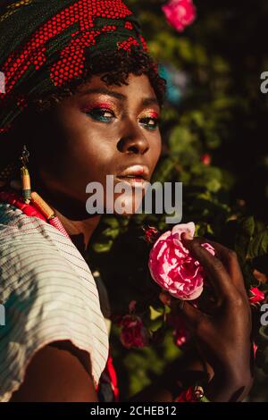 Zugeschnittenes Bild der afrikanischen weiblichen Modelin Nationalkostüm, Schmuck, Turban und Make-up posiert im Garten von Rosen, hellen Tageslicht Sonne. Stockfoto