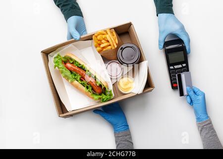 Die Hände des Kuriers in den Handschuhen, neben der Schachtel mit dem Hot Dog, dem Kaffee in der Tasse, den Kartoffeln, dem Ketchup und dem Senf im Behälter Stockfoto