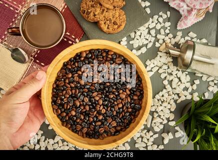 Mischung aus natürlichem und geröstetem Kaffee in einer Schüssel, die von einer kaukasischen Hand auf einem Holztisch mit Keksen, einer Tasse Kaffee, einem Löffel und Zucker gehalten wird Stockfoto