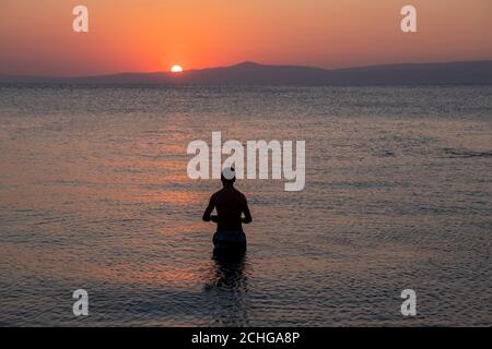 Orangefarbene Sonne untergeht im Morgengrauen hinter den Bergen. Schwarze Silhouette eines jungen Mannes, der in farbenfroh blauem Wasser steht und von kleinen Wellen umgeben ist Stockfoto