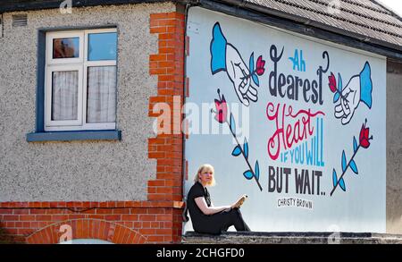 Die Künstlerin Sarah Bracken Soper liest unter einem Wandgemälde, das sie auf der Seite ihres Hauses in Crumlin Dublin während der Covid-19 Lockdown malte, eine Kopie von My Left Foot von Christy Brown. Stockfoto