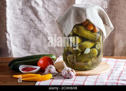 Eingelegte Gurken und Tomaten in einem Glas auf Leinentischdecke und Holztisch. Selbstgemacht, Kopierbereich. Stockfoto