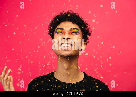 Fröhlicher Mann mit Regenbogen Auge Make-up werfen den Glitter. Aufgeregt Geschlecht Flüssigkeit männlich Blick auf fallen glitzert vor rotem Hintergrund. Stockfoto