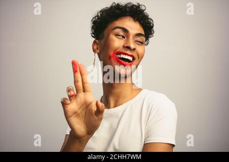 Lächelnder schwuler Mann mit verschmierter Lippenstift. Junger Mann mit verschmiertem Lippenstift auf seinem Gesicht vor weißem Hintergrund. Stockfoto