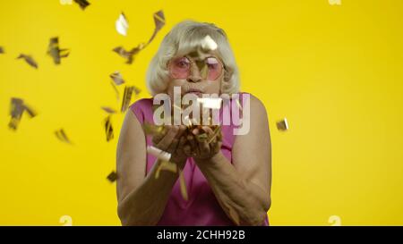 Glückliche ältere alte Frau lacht, weht Konfetti glitzert, Geburtstag gratuliert zum Jahrestag, Lotterie zu gewinnen. Ältere stilvolle Dame Oma auf gelbem Hintergrund Stockfoto