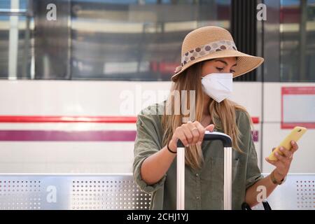 Junge Frau trägt Gesichtsmaske und Explorer Hut, mit ihrem Smartphone, während auf den Zug an einem Bahnhof wartet. Reisekonzept. Stockfoto