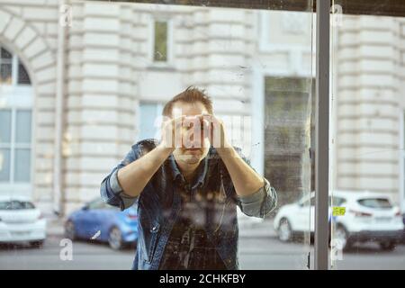 Der junge Mann schaut durch das Glas des öffentlichen Nahverkehrs in der Innenstadt. Kaukasischer Mann auf der Straße in der Nähe der Bushaltestelle. Stockfoto
