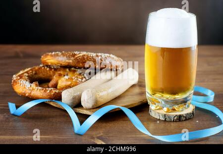 Stillleben mit einem Becher leichten Bieres, Brezeln und weiß gekochten bayerischen Würsten auf dem Holz. Leckere und typische Snacks und Getränke am Tag des Oktoberfests Stockfoto