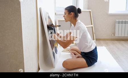 Konzentrierte Brünette mit Brötchen in weißem T-Shirt und schwarzen Shorts Zeichnet Bild laufende Hände mit Farben auf Leinwand in der Werkstatt Im Sommer Stockfoto