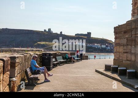 Die Leute saßen entspannt auf Bänken auf dem Pier in Whitby, North Yorkshire, England, Großbritannien mit der Abtei und der Kirche im Hintergrund Stockfoto