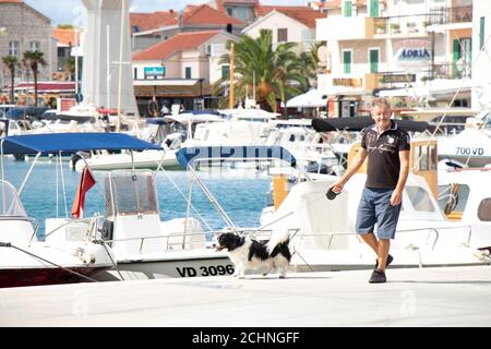 Vodice, Kroatien - 1. September 2020: Mann, der einen Hund an der Leine auf dem malerischen Dock mit Booten und Häusern läuft Stockfoto