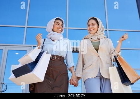 Low-Angle-Porträt von zwei jungen Frauen aus dem Nahen Osten beim Einkaufen Taschen und lächeln einander an, während Sie Mall mit verlassen Käufe Stockfoto