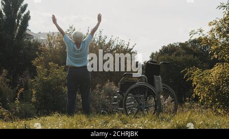 Ältere Frau mit ausgestreckten Händen in der Natur vor dem Rollstuhl. Hochwertige Fotos Stockfoto