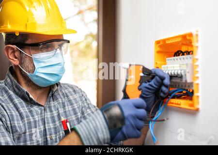 Elektriker bei der Arbeit an einem elektrischen Panel mit Helm, Schutzbrille und Handschuhe geschützt; tragen Sie die chirurgische Maske, um die Ausbreitung von Coronavirus zu verhindern Stockfoto