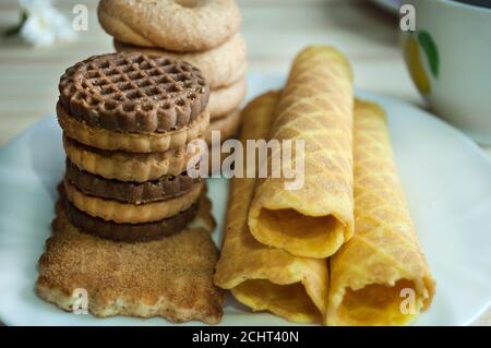 Waffelbrötchen und Kekse auf einem weißen Teller und eine Tasse Tee. Waffeln auf der Seite. Komposition auf einem hölzernen Hintergrund. Sichtbare Waffelstruktur Stockfoto