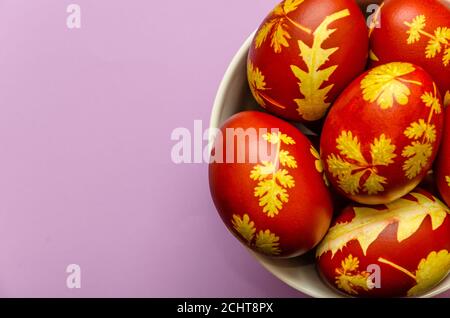 Ostereier mit Zwiebelschalen gefärbt mit einem Muster aus frischen Pflanzenblättern in einer weißen Schüssel auf hellviolettem Hintergrund. Konzept der Verwendung von natürlichen Farbstoffen Stockfoto