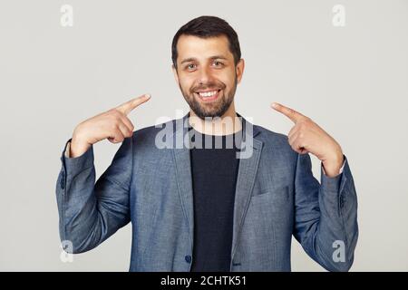 Junger Geschäftsmann Mann mit Bart in Jacke lächelnd selbstbewusst zeigen und zeigen mit den Fingern Zähne und Mund. Gesundheitskonzept. Porträt eines Mannes auf grauem Hintergrund. Stockfoto