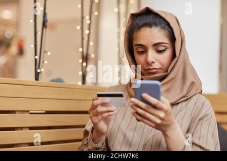 Porträt einer jungen Frau aus dem Nahen Osten, die Kopftuch trägt und Online-Einkäufe über das Smartphone macht, während sie im Café sitzt und mit Kreditkarte und Kopierraum hält Stockfoto