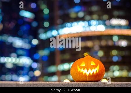 Halloween Kürbis auf dem Hintergrund der Nachtstadt. Mehrstöckige Gebäude und Wolkenkratzer. Verschwommene farbige Lichter der Stadtbeleuchtung. Nachtstadt Stockfoto