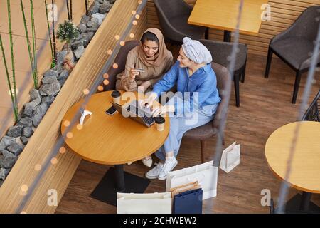 Blick aus dem hohen Winkel auf zwei junge Frauen aus dem Nahen Osten, die online per Laptop einkaufen, während sie im Café eines Einkaufszentrums sitzen und mit Kreditkarte und Kopierplatz arbeiten Stockfoto