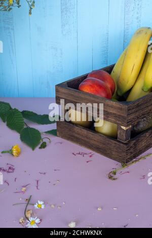 Obstkiste aus Holz mit Banane, Orangen, Kiwi, Pfirsich, Aprikose und Blumen auf rosafarbenem Tisch mit blauem Hintergrund Stockfoto
