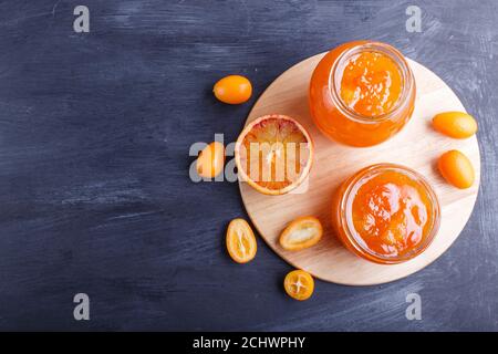 Orangen- und Kumquat-Marmelade in einem Glas mit frischen Früchten auf schwarzem Holzgrund. Selbstgemacht, Draufsicht, Kopierbereich. Stockfoto