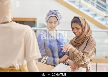 Waist-up-Porträt von zwei jungen Frauen aus dem Nahen Osten, die auf Schaufenster zeigen, während Schaufensterbummel in der Mall, Copy Space Stockfoto