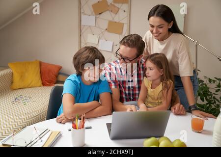 Junge glückliche Familie, die zusammen am Laptop arbeitet, Eltern helfen Kindern bei Hausaufgaben oder studieren online zu Hause während covid 19 Quarantäne Stockfoto