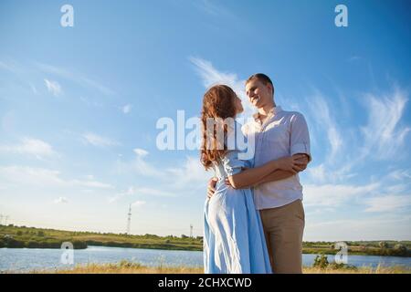 Ein junges Paar umarmt sich auf einem Feld vor dem Hintergrund eines Flusses oder Sees und genießt das Leben. Das Konzept der Liebe und korrekte nicht-missbräuchliche Beziehungen. Stockfoto