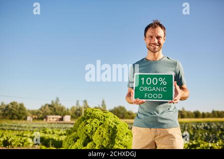 Porträt eines lächelnden Mannes, der ein Schild MIT BIO-LEBENSMITTELN hält und die Kamera anschaut, während er im Sonnenlicht auf der Gemüseplantage steht, Raum kopieren Stockfoto