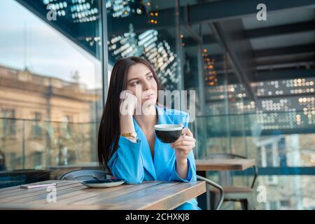 Nachdenkliche Frau auf dem Balkon oder der trendigen Terrasse im Coffeeshop, die eine Tasse heißes Getränk serviert. Junge schöne Modell genießen Mittagessen Zeit Tasse Kaffee, t Stockfoto