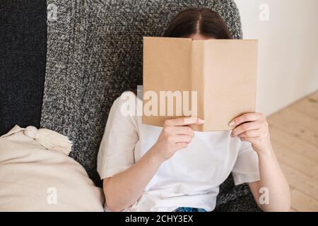 Junge Frau in Jeans und weißem T-Shirt sitzt in grauem Sessel und liest Papierbuch. Konzept von erholsamen Urlaub, Wohnkomfort, Hygge, Slow-Living Stockfoto