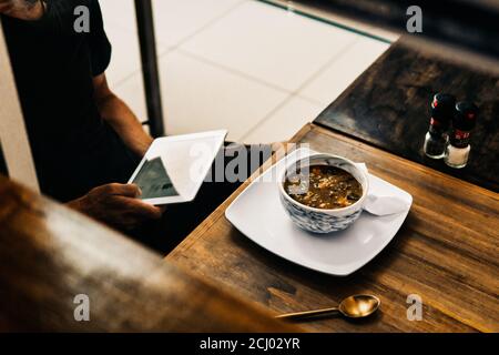 Geschäftsmann, der während seiner Mittagspause mit auf sein Tablet schaut Eine Suppe auf dem Tisch Stockfoto