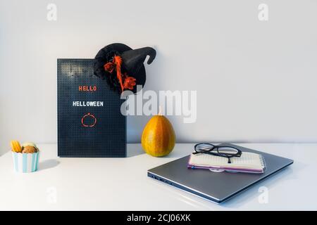 Minimalistisches Halloween Dekor - Brieftafel mit Hello Halloween Nachricht, Hexenhut, dekorativer Kürbis, und Tasse mit süßen Makronen auf weißem Arbeits t Stockfoto